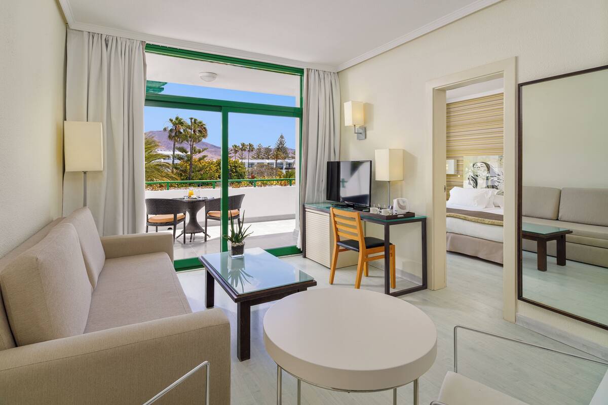 A hotel room with a sofa, a small round table, a TV, and a desk with a chair. There is a balcony with a view of palm trees and mountains.
