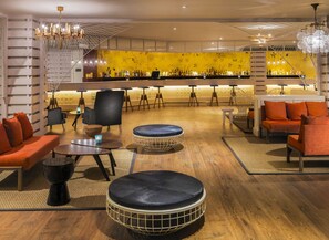 A modern bar area with a yellow wall, wooden flooring, and various seating options including sofas and round tables. - H10 Lanzarote Princess (Yaiza)