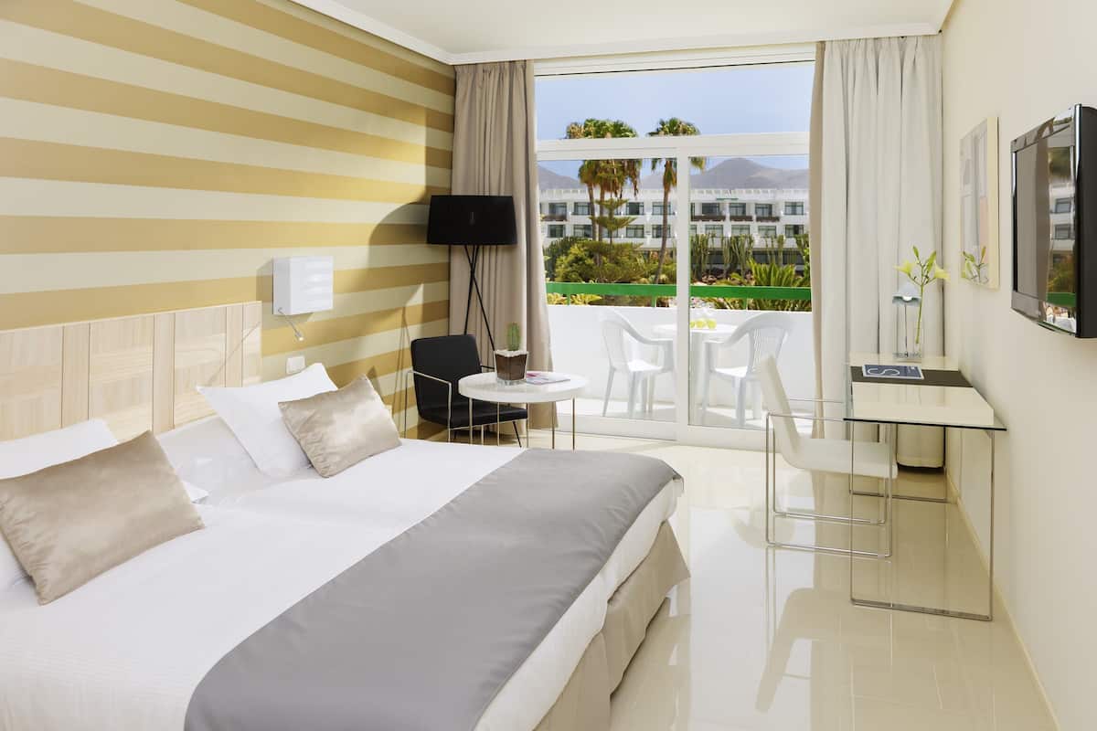 A hotel room with a bed, a desk, a TV, and a balcony with outdoor seating.