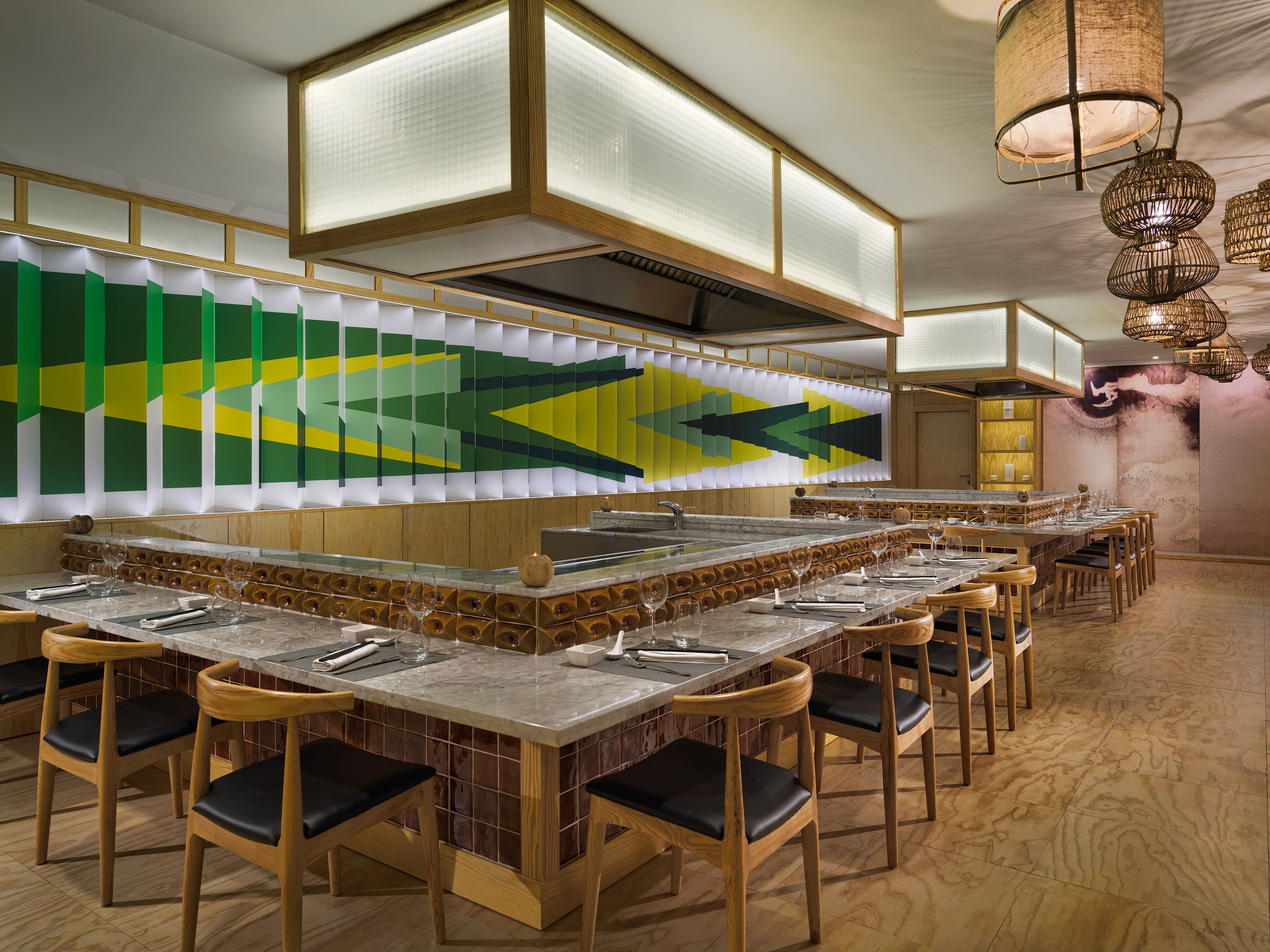 Un restaurante con una barra larga, varias sillas y una pared decorativa con patrones geométricos.