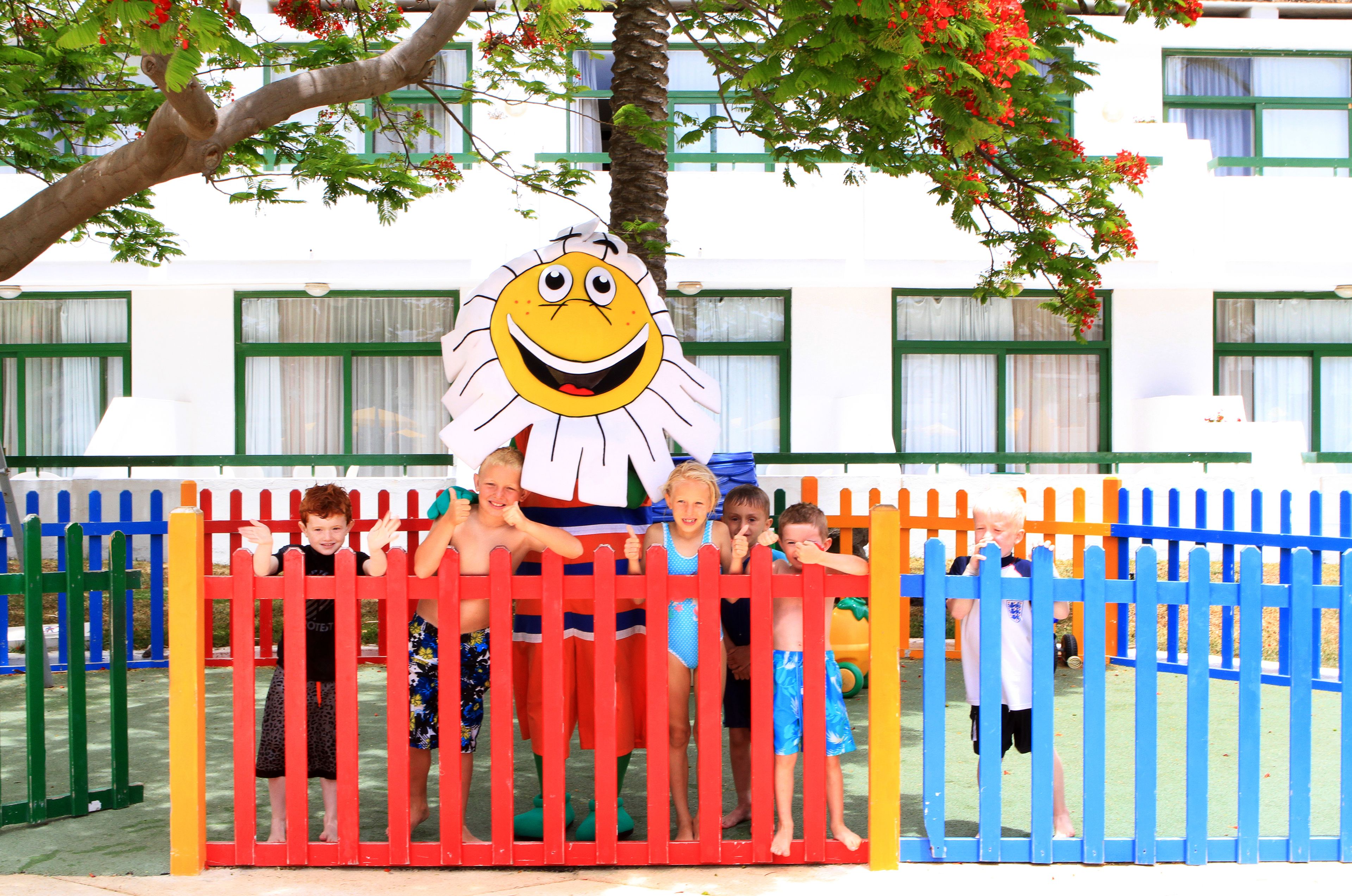 A group of children playing behind a colorful fence with a large cartoon character in the background.