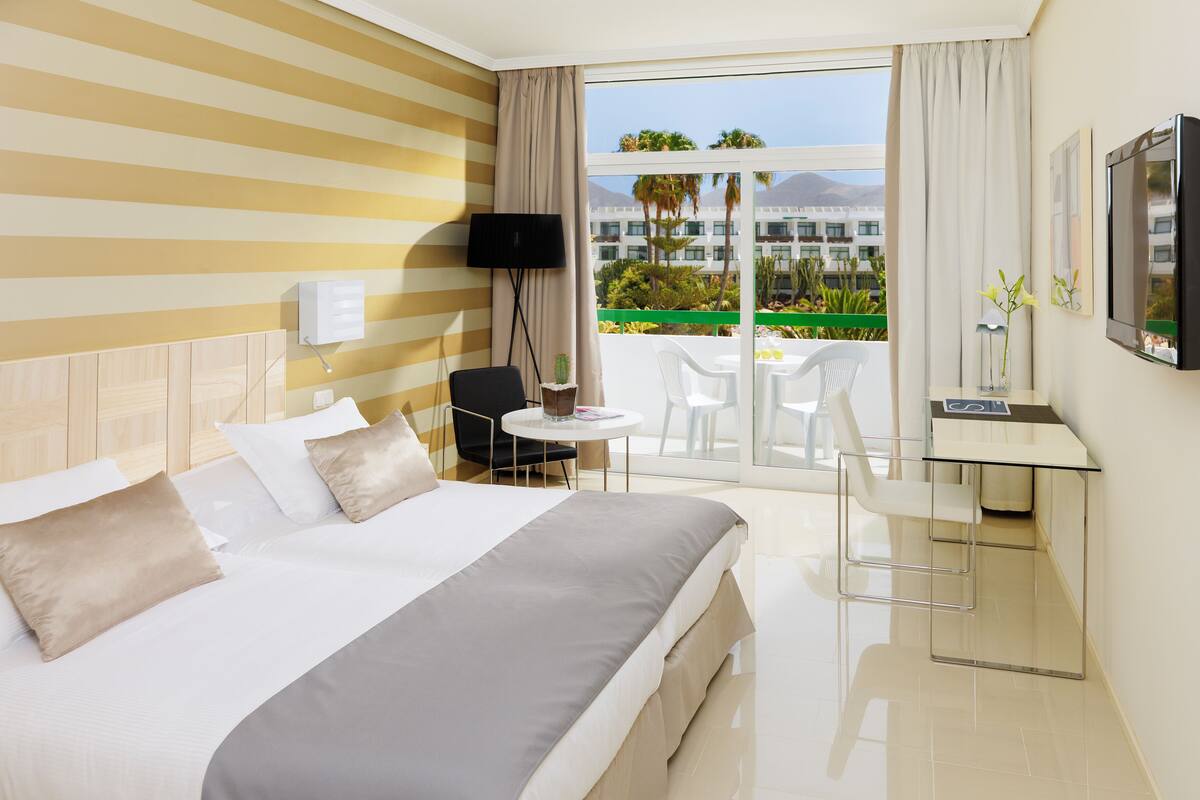 A hotel room with a bed, a desk, a TV, and a balcony with a view of palm trees and buildings.