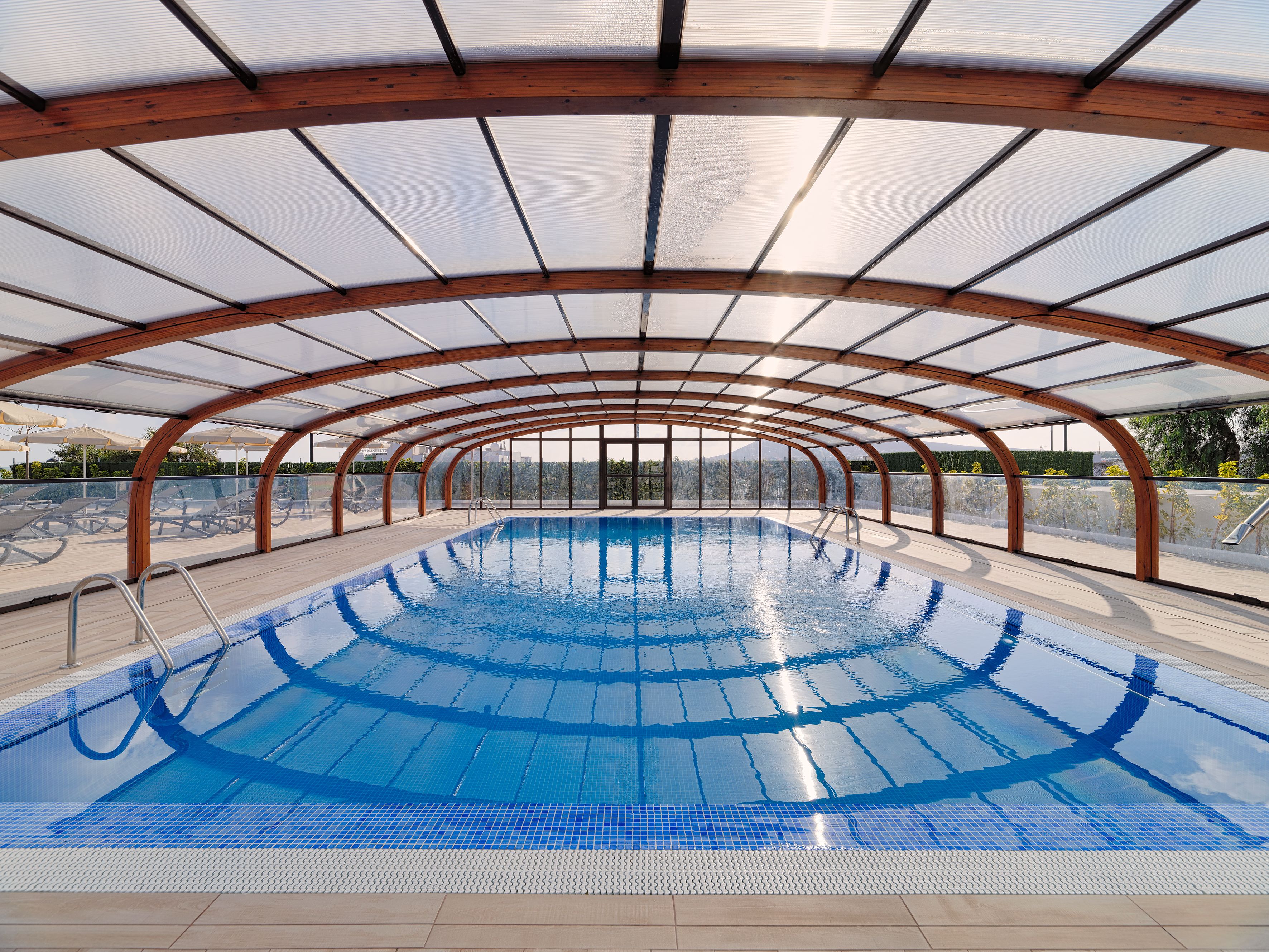 A covered swimming pool with a wooden frame and glass panels, surrounded by lounge chairs and a view of the cityscape.