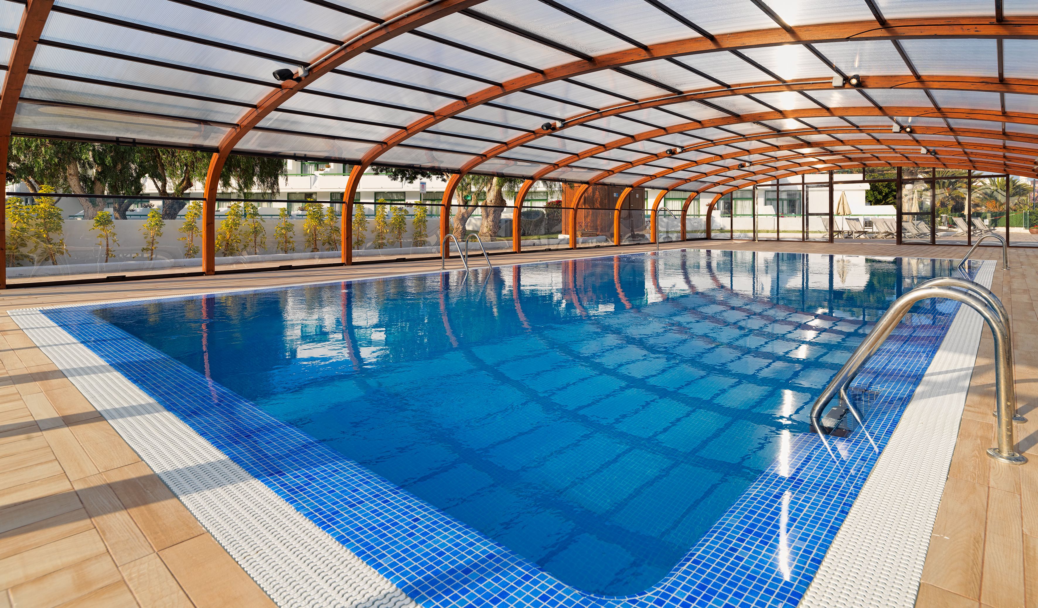 Una piscina interior con agua cristalina azul, rodeada por una cubierta de madera y una estructura de techo de vidrio.