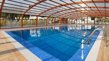 An indoor swimming pool with a clear blue water, surrounded by a wooden deck and a glass ceiling structure.