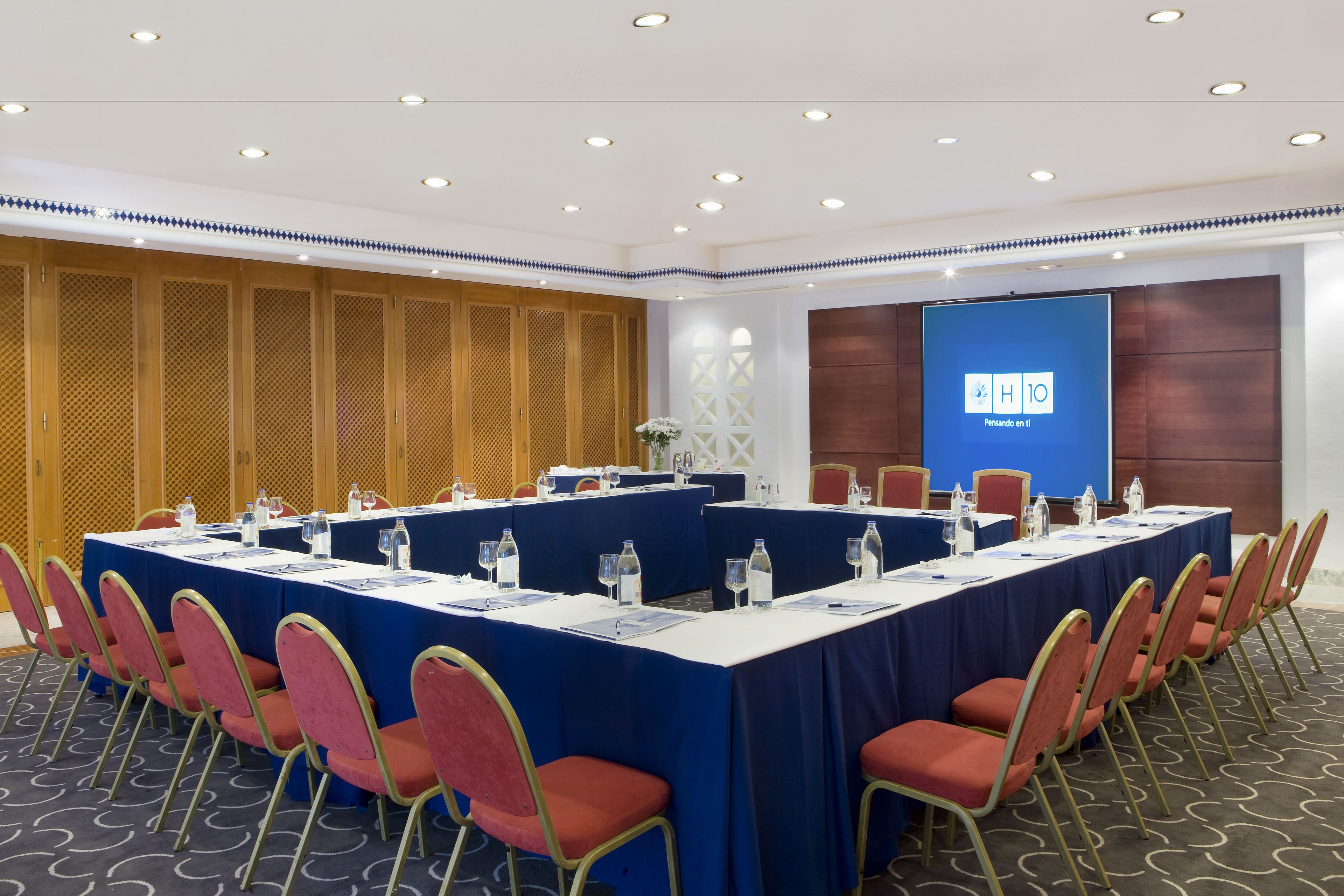 Un salón de conferencias con una larga mesa, sillas rojas y una pantalla azul que muestra 
