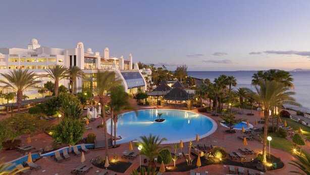 A resort with a large pool, palm trees, and a view of the ocean. - H10 Timanfaya Palace (Yaiza)