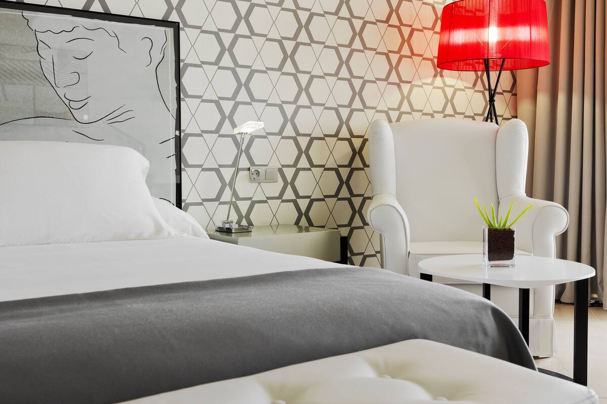 Una moderna camera d'albergo con una parete a motivi geometrici, un letto bianco, una lampada rossa, un tavolino con una pianta e una poltrona bianca.