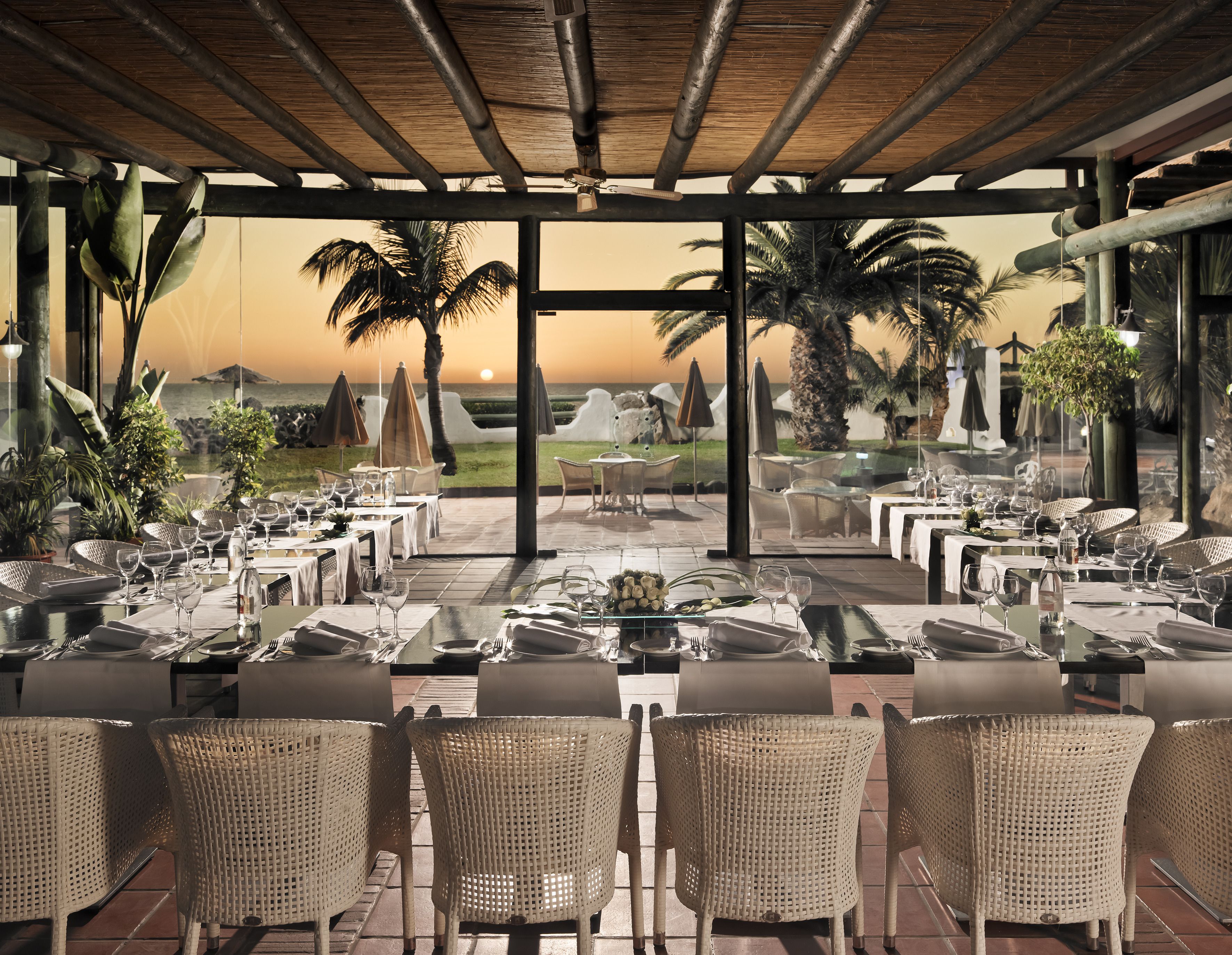 Un área de comedor al aire libre con mesas y sillas, con vista a una playa y palmeras.