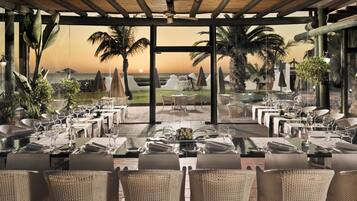 Un área de comedor al aire libre con mesas y sillas, con vista a una playa y palmeras.