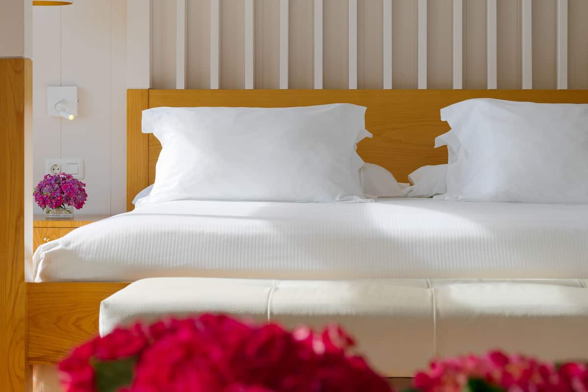 A neatly made bed with white linens, a wooden headboard, and a bedside table with a vase of pink flowers.