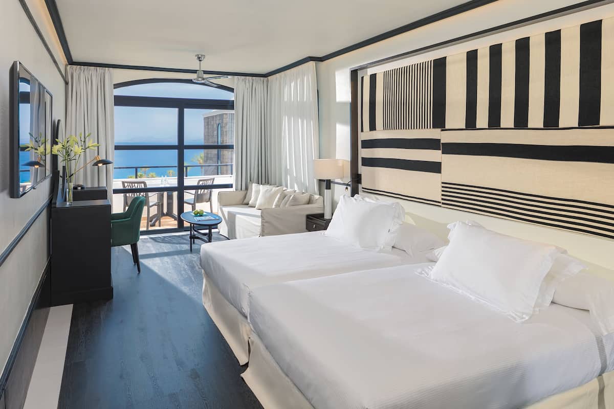 A hotel room with two beds, a desk, a chair, a TV, and a balcony with a view of the sea.