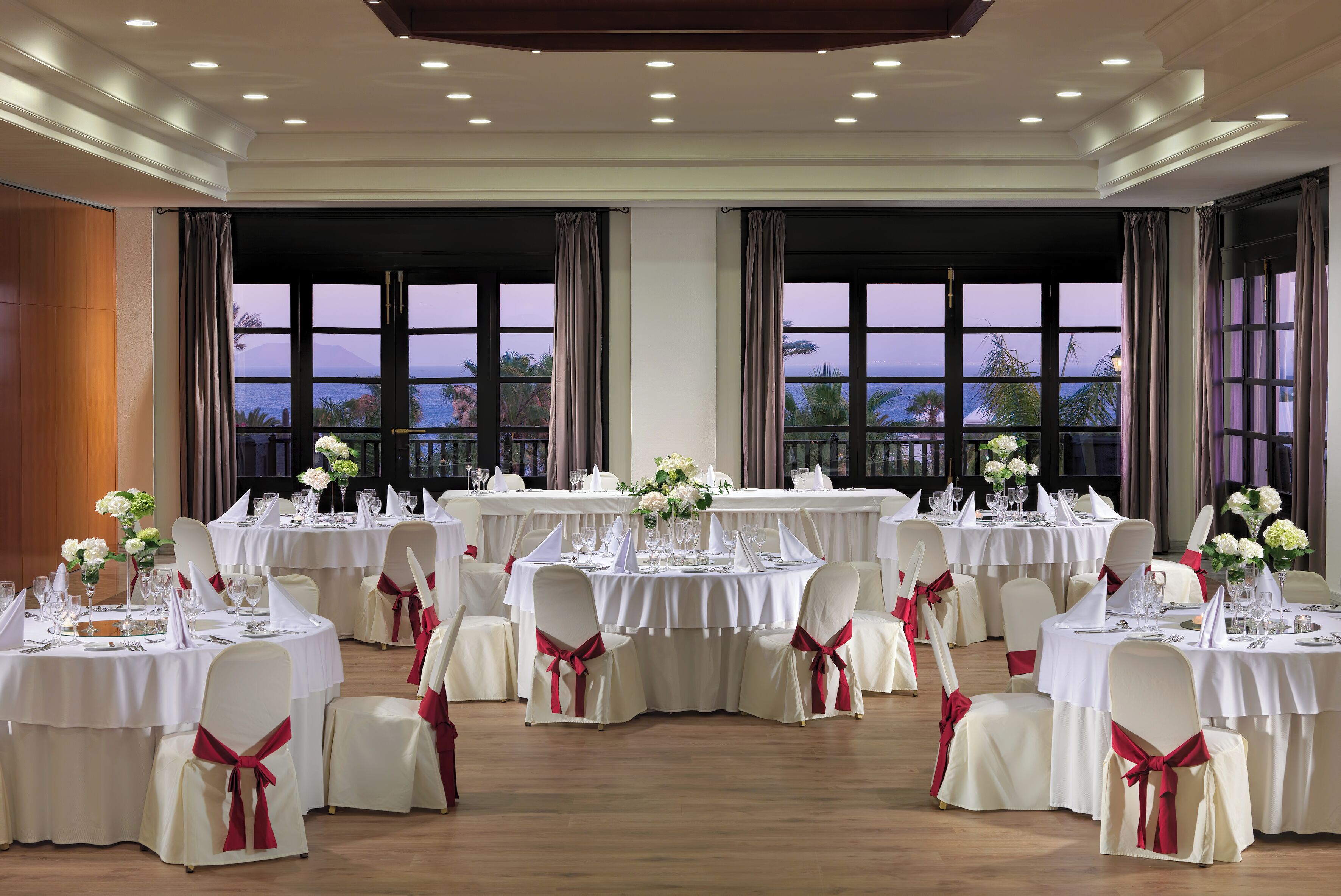 Una sala da banchetti con diversi tavoli rotondi apparecchiati per la cena, ampie finestre con tende e vista sul mare.