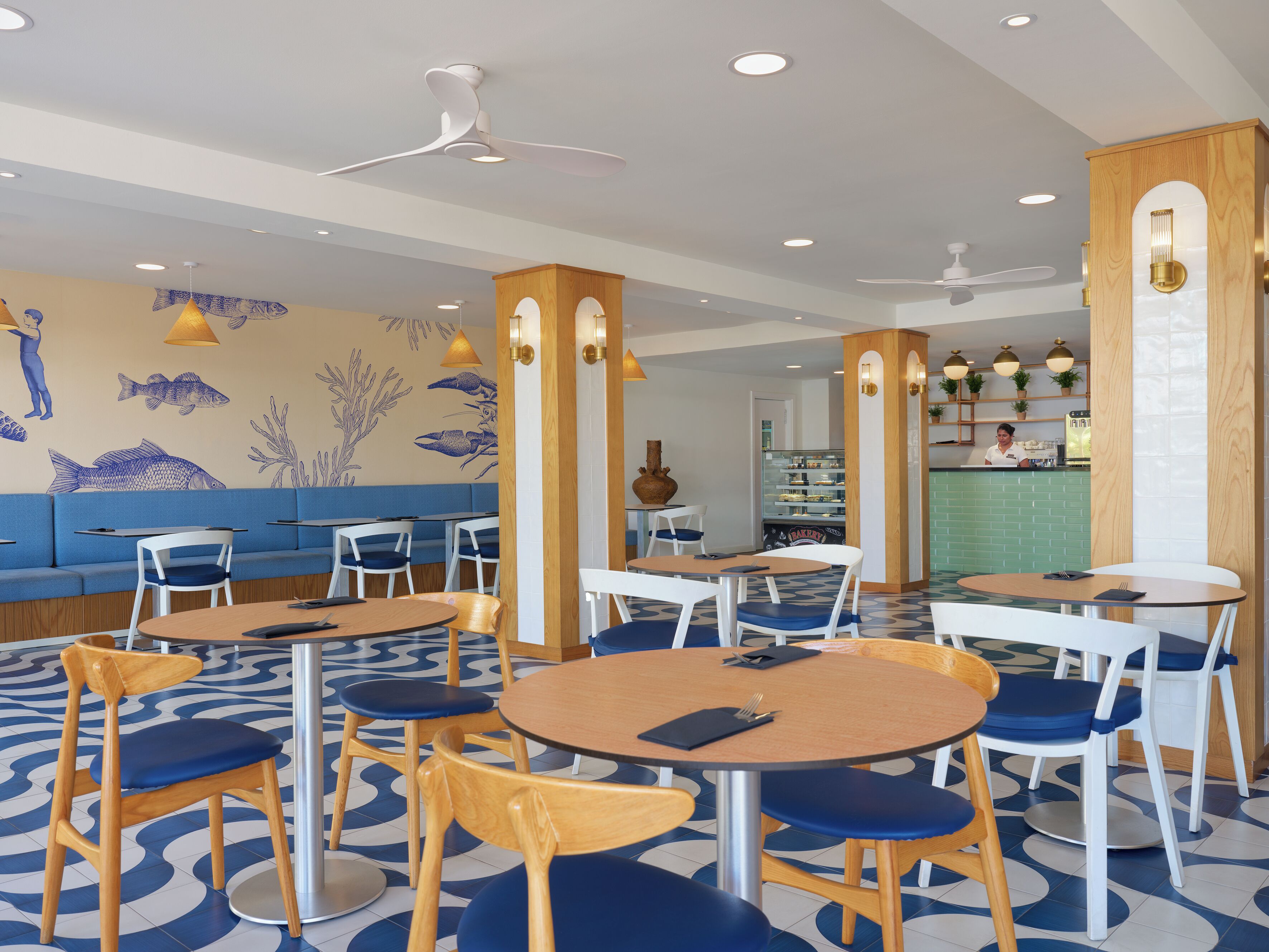 A restaurant interior with round tables, blue cushioned chairs, a blue and white patterned floor, and wall murals of fish and sea plants.