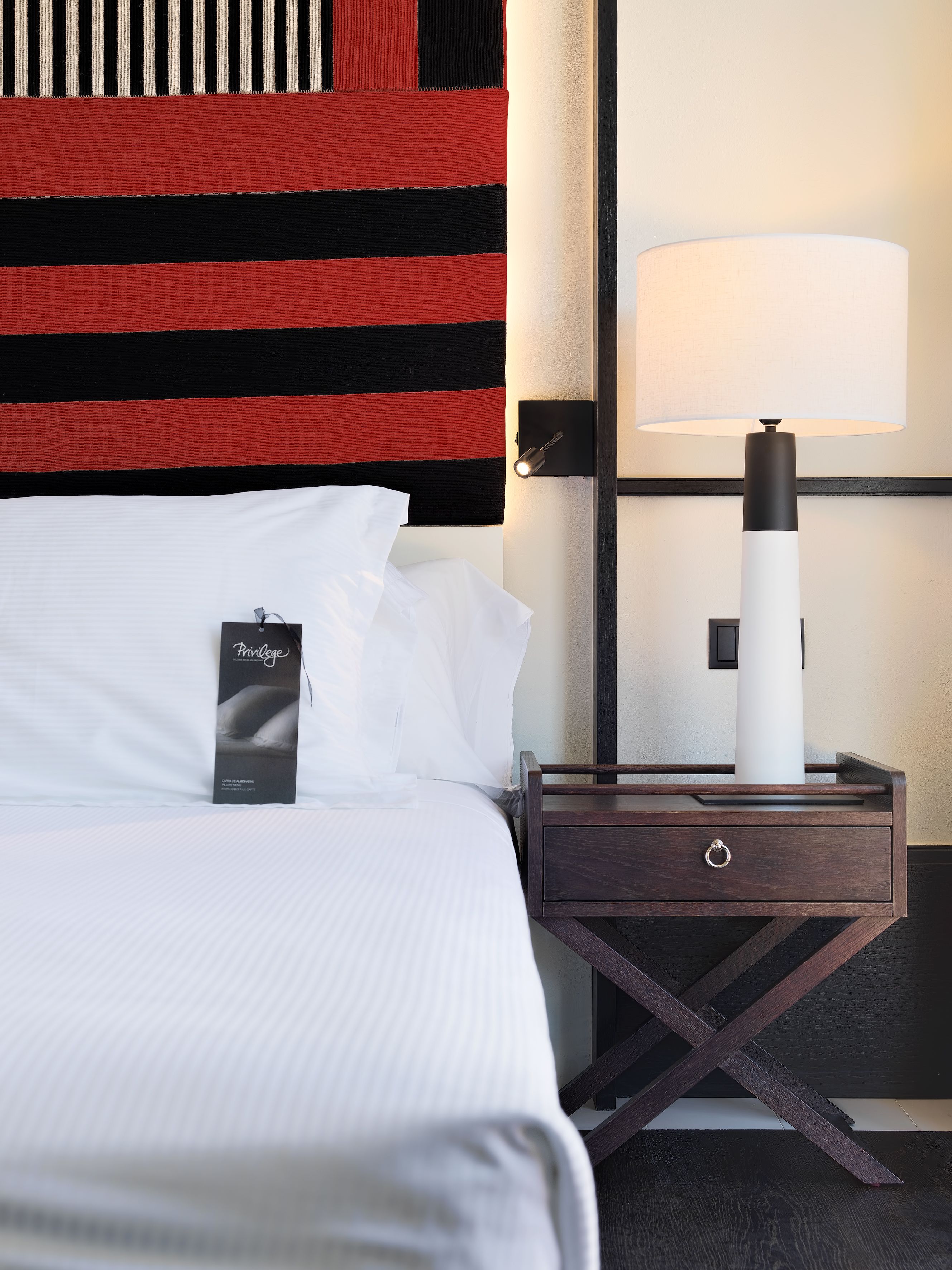 A neatly made bed with white linens, a black tag on the pillow, a dark wooden nightstand with a lamp, and a striped wall behind the bed.
