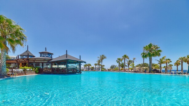 A large, clear blue pool with lounge chairs and umbrellas, surrounded by palm trees and a building with a gazebo.