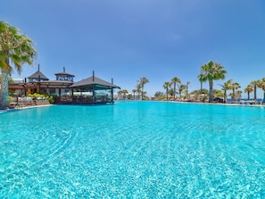 A large, clear blue pool with lounge chairs and umbrellas, surrounded by palm trees and a building with a gazebo. - H10 Rubicón Horizons Collection (Yaiza)