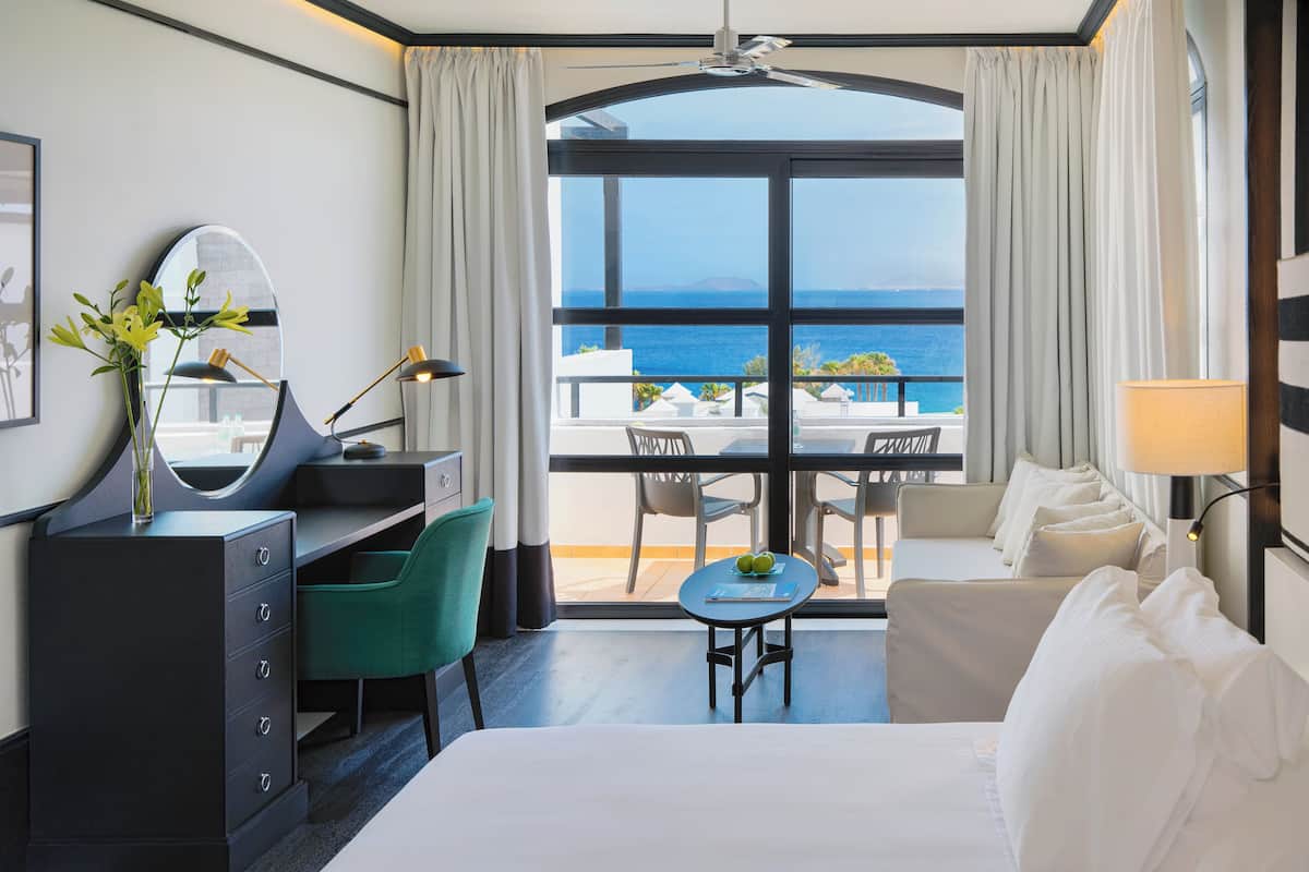 A hotel room with a bed, a desk, a chair, a small table, and a view of the sea.