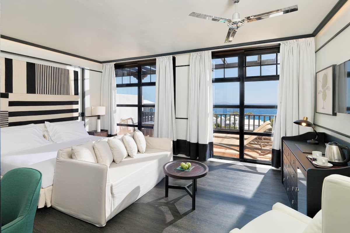 A modern hotel room with a bed, sofa, and a balcony with a view of the sea.