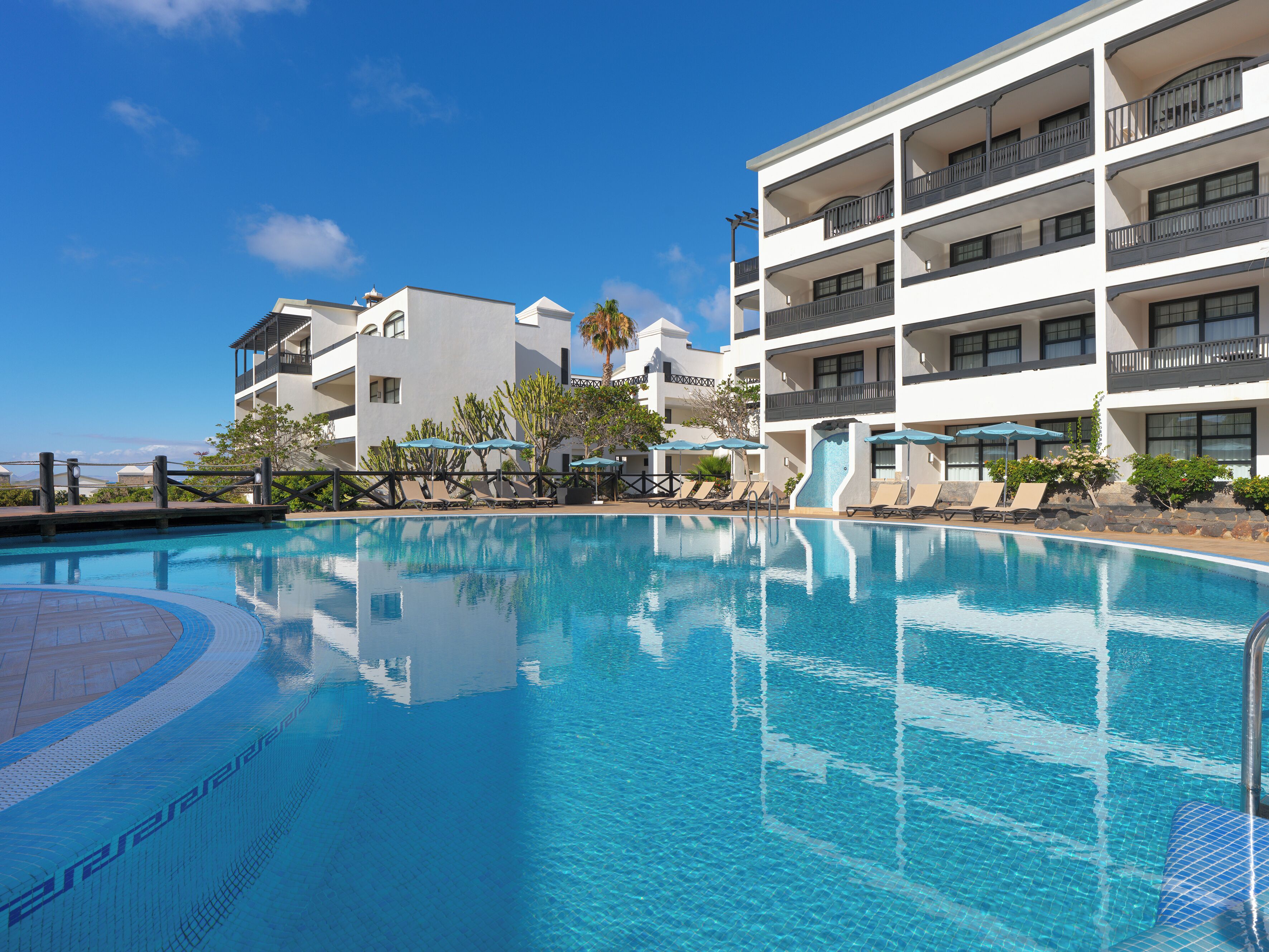 Une piscine entourée de chaises longues et de parasols, elle-même bordée d’un immeuble à plusieurs étages doté de balcons.