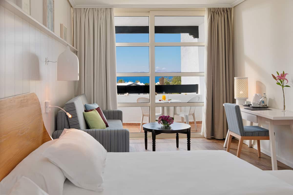 A hotel room with a bed, a sofa, a dining table with chairs, a desk with a chair, and a large window with a view of the sea.