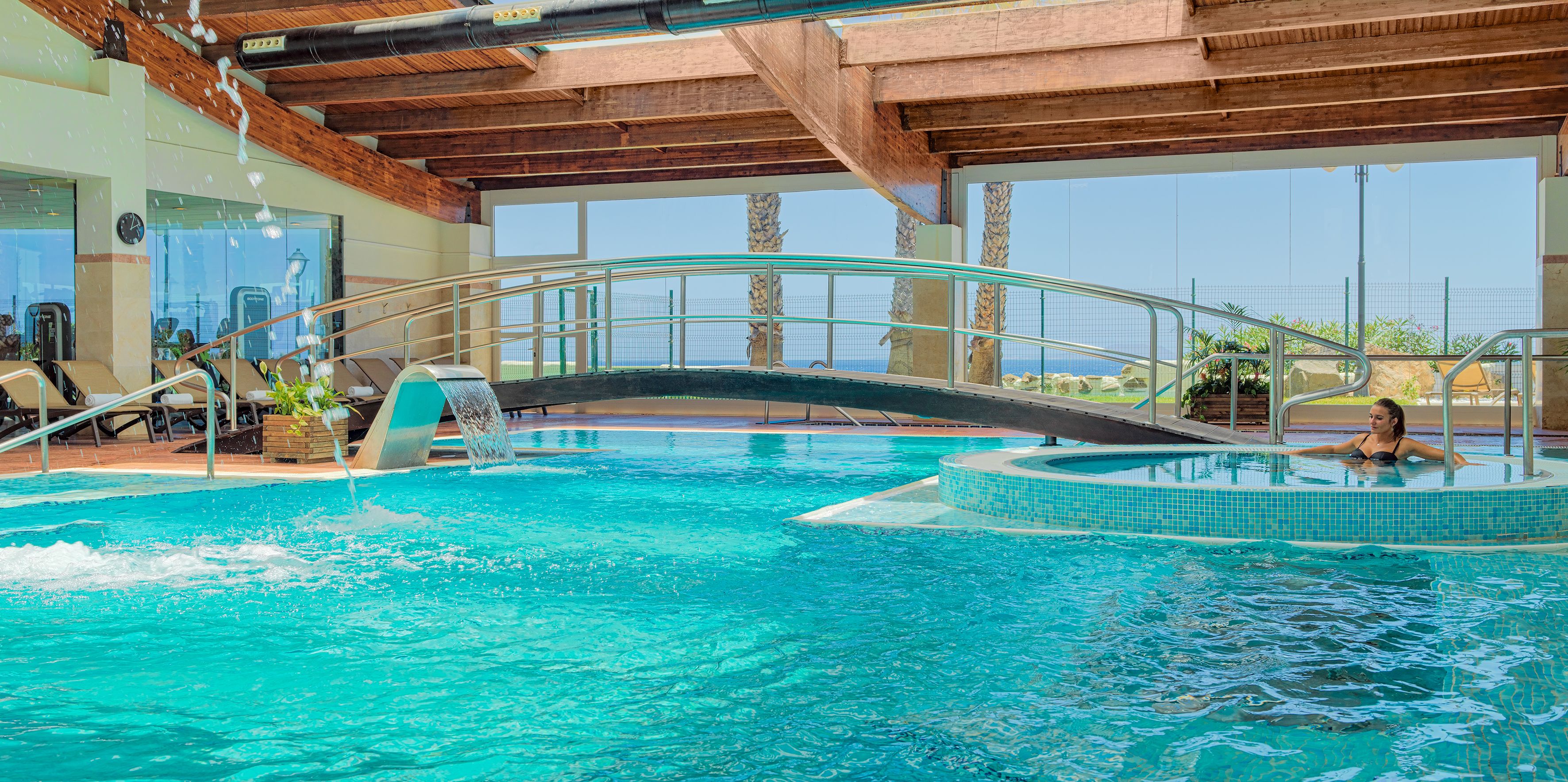 Una piscina interior con un puente de cristal, sillas de descanso y una bañera de hidromasaje.