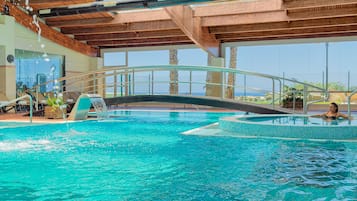 Una piscina interior con un puente de cristal, sillas de descanso y una bañera de hidromasaje.