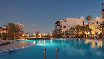 Una piscina con sillas de descanso y una terraza de madera, rodeada de edificios y palmeras.