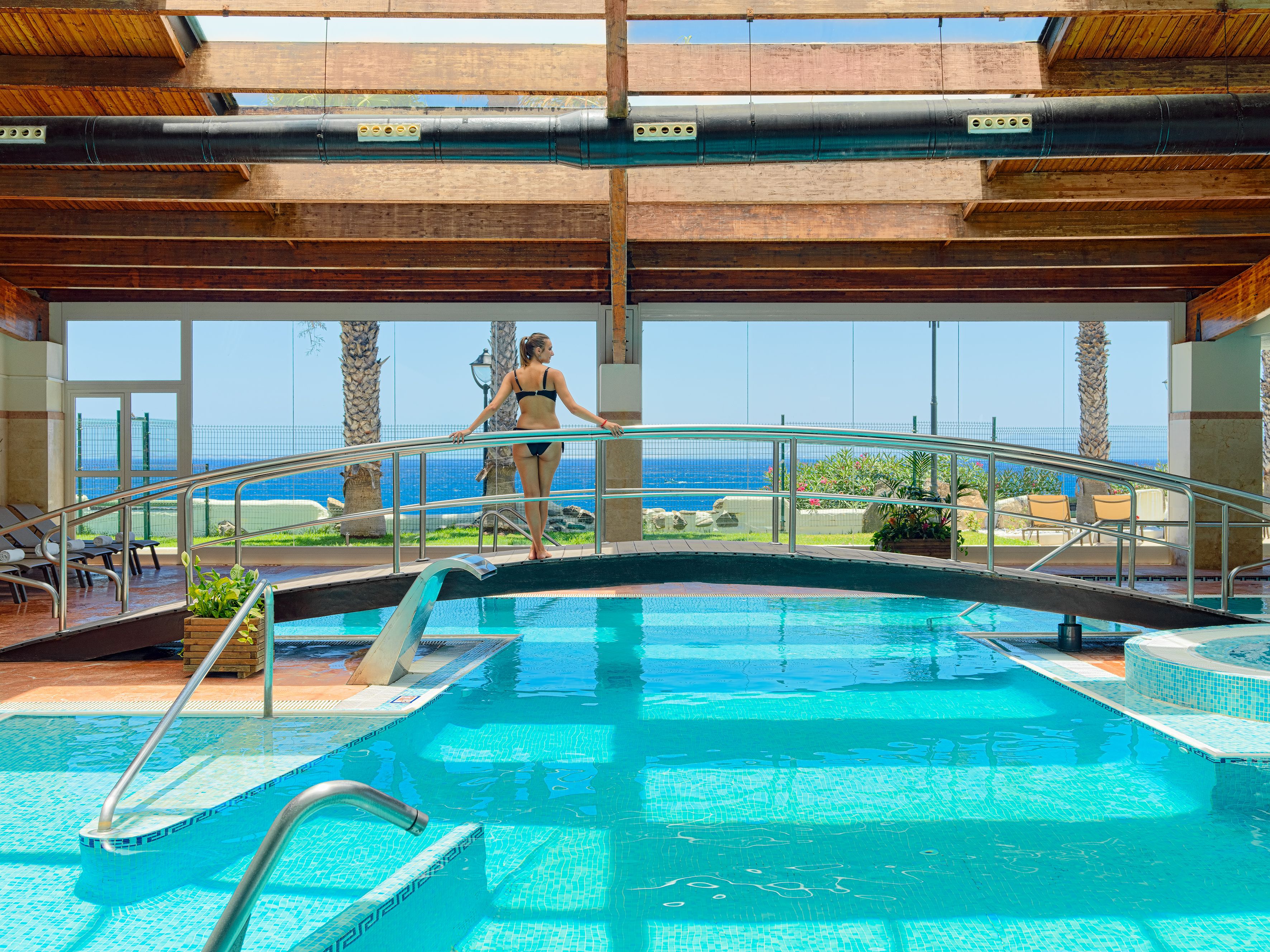 Piscina com vista para o oceano, uma mulher de biquíni e estrutura de teto de madeira.
