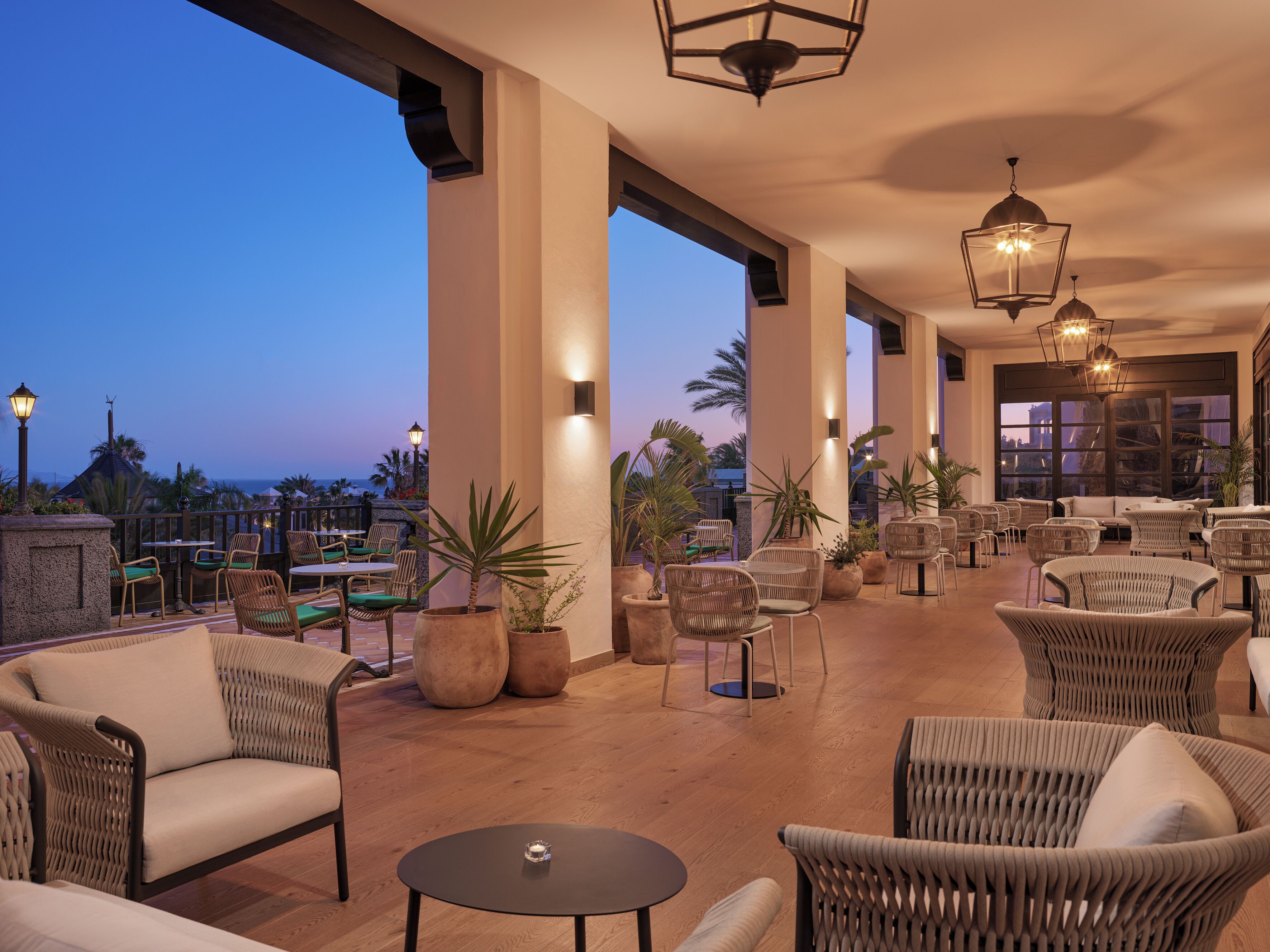 A spacious outdoor seating area with wicker furniture, potted plants, and a view of the ocean at dusk.