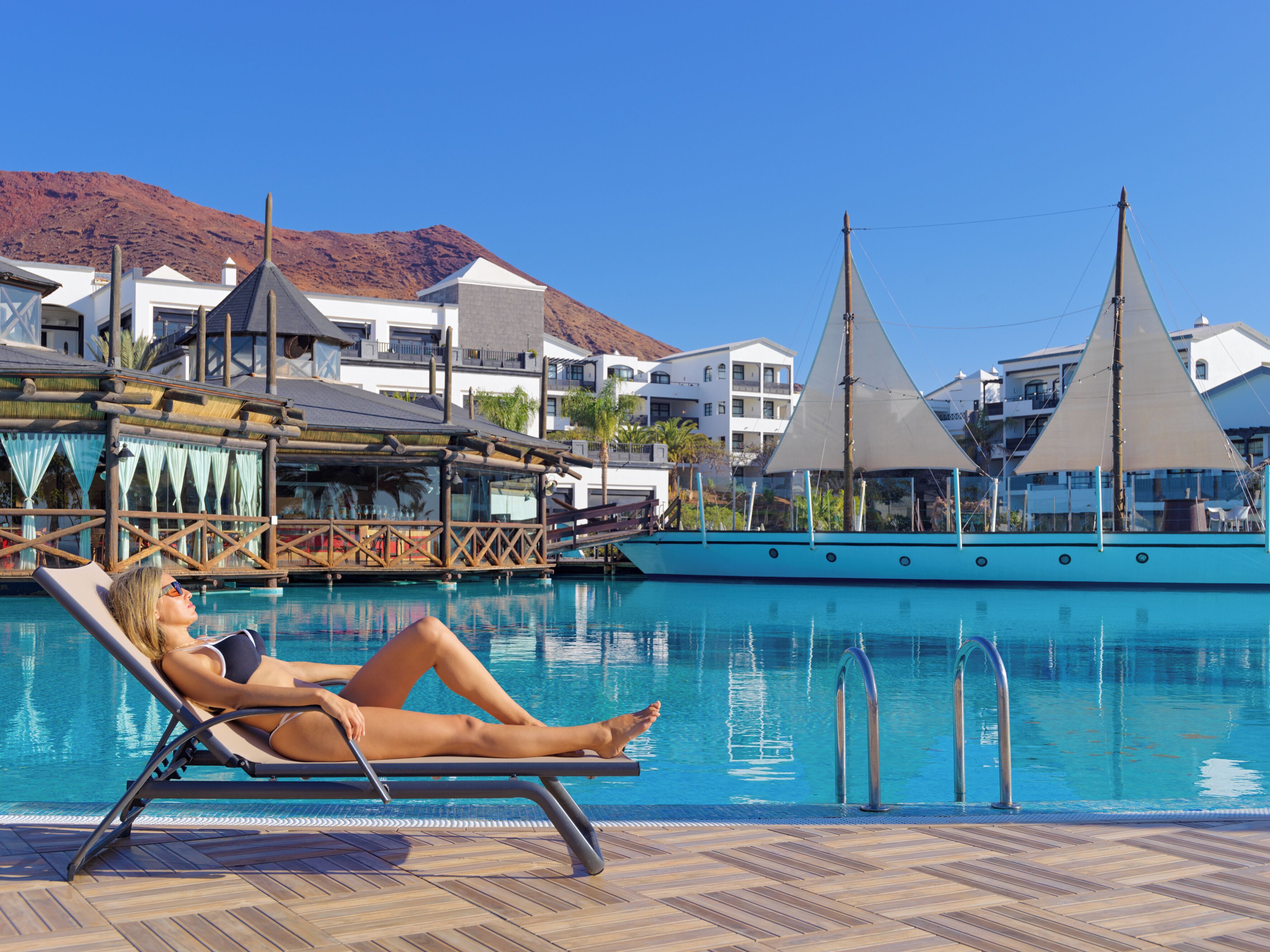 Zona de piscina con una silla de descanso, un bote y un edificio con montañas al fondo.
