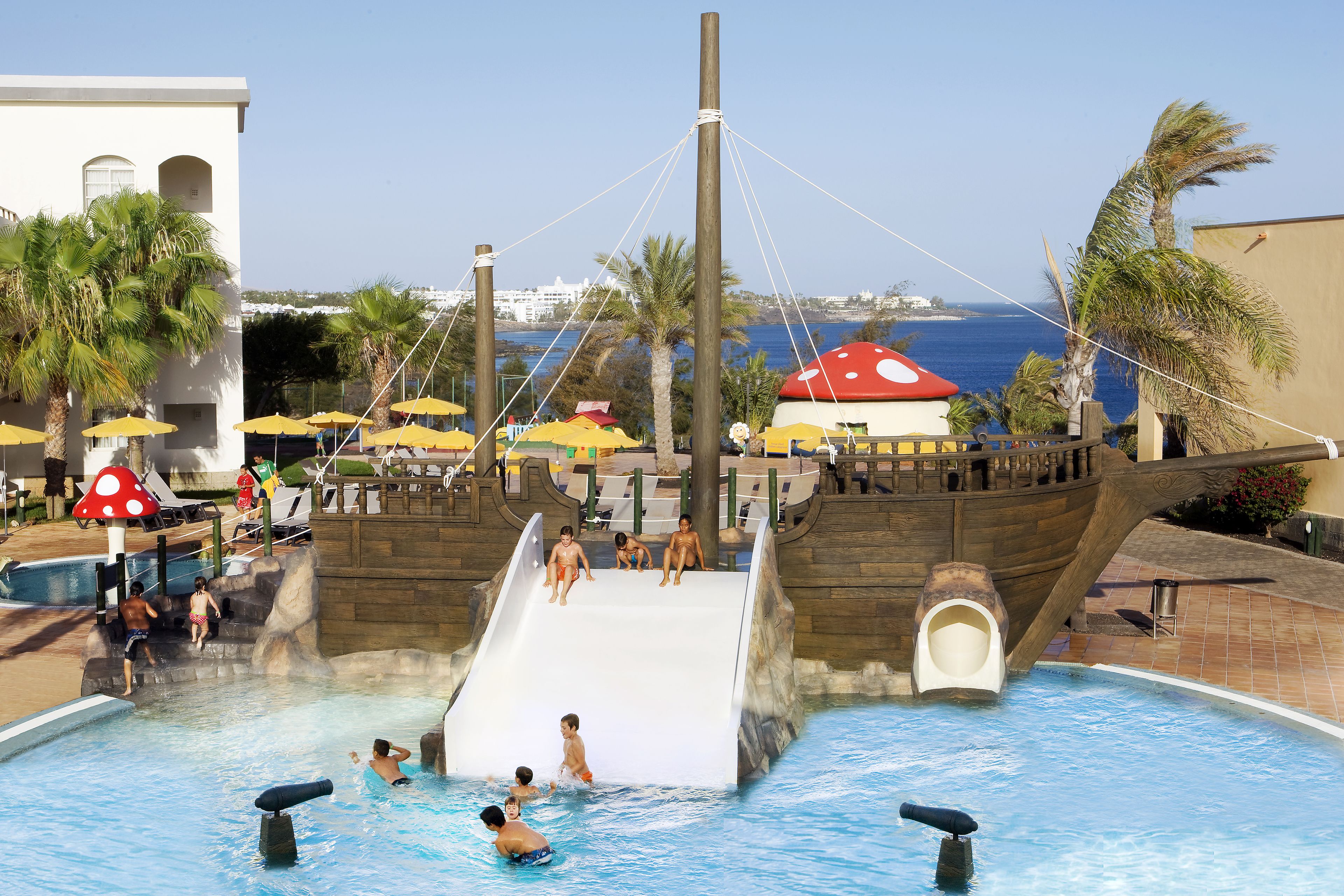 A pool area with a pirate ship slide, palm trees, and umbrellas.