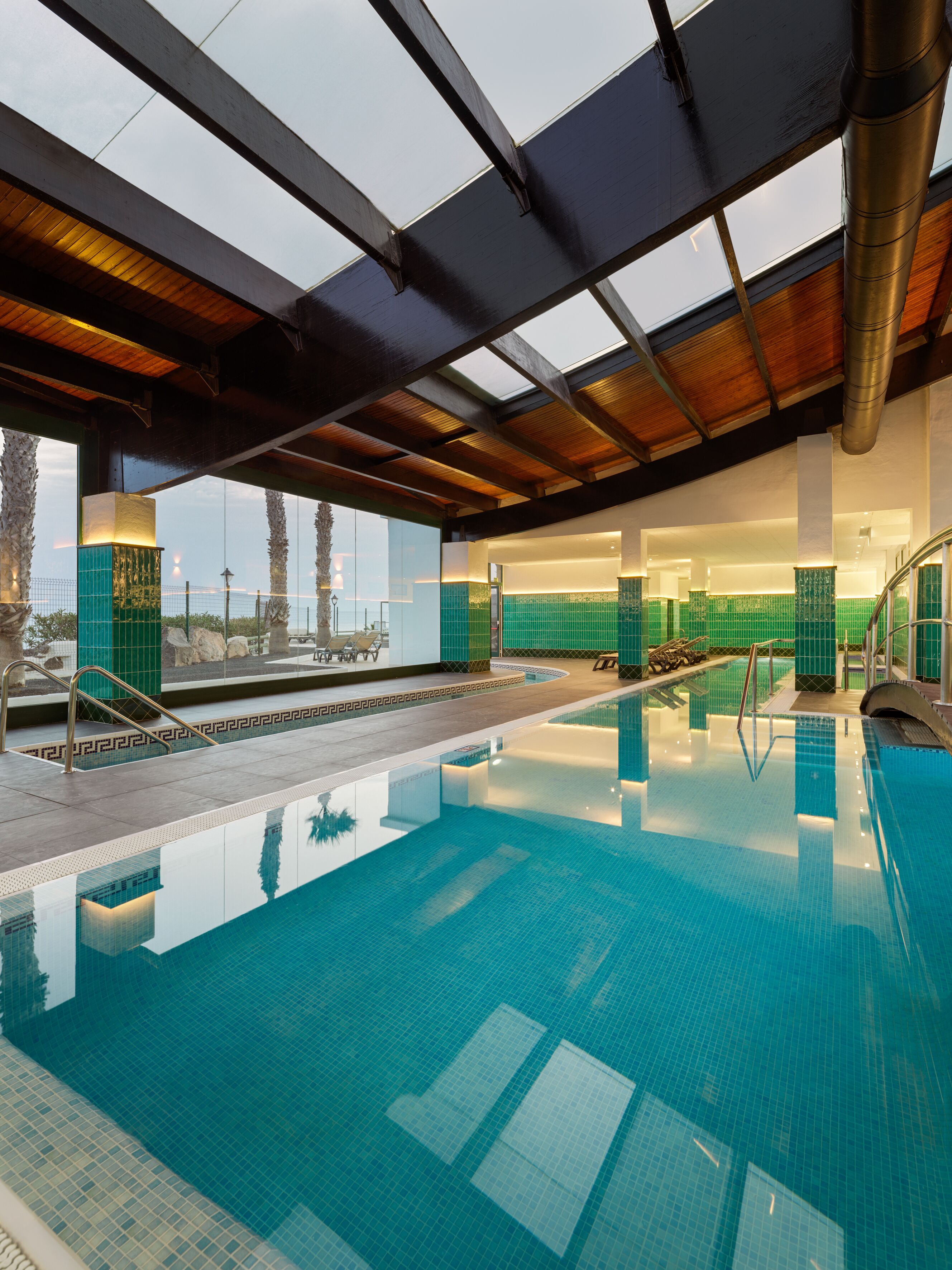 An indoor swimming pool with clear blue water, a wooden ceiling, and a view of the ocean through large windows.