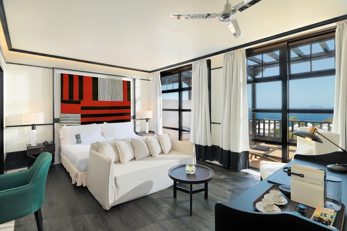 A hotel room with a bed, a sofa, a coffee table, a desk, and a view of the sea.