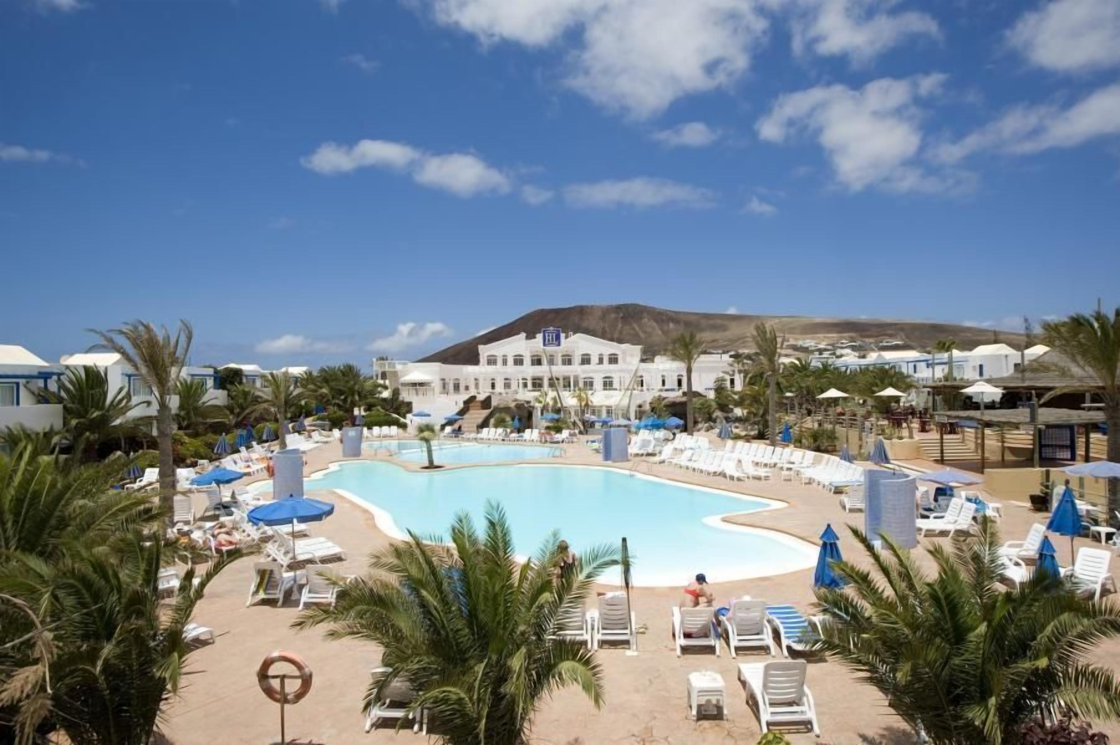 Un complexe hôtelier doté d’une grande piscine entourée de transats et de parasols, avec une montagne en arrière-plan et un bâtiment portant une enseigne.