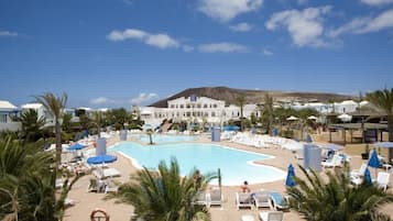 Un resort con una gran piscina rodeada de tumbonas y sombrillas, una montaña al fondo y un edificio con un letrero.