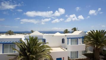Un hotel frente al mar con edificios blancos y detalles azules, con palmeras y vistas al océano y las montañas.