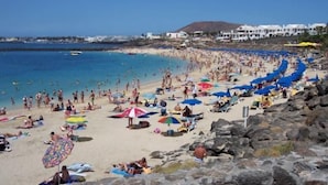 Une plage bondée où les gens se prélassent au soleil et se baignent, un rivage rocheux et des bâtiments en arrière-plan.