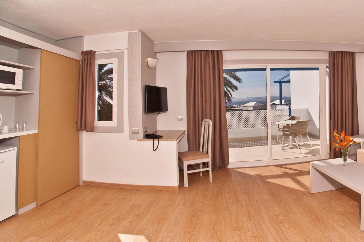 Sala de estar moderna com sofá, mesa de centro, TV, escrivaninha pequena e uma cadeira. Há uma porta de vidro deslizante que dá para uma varanda com vista para palmeiras e prédios.