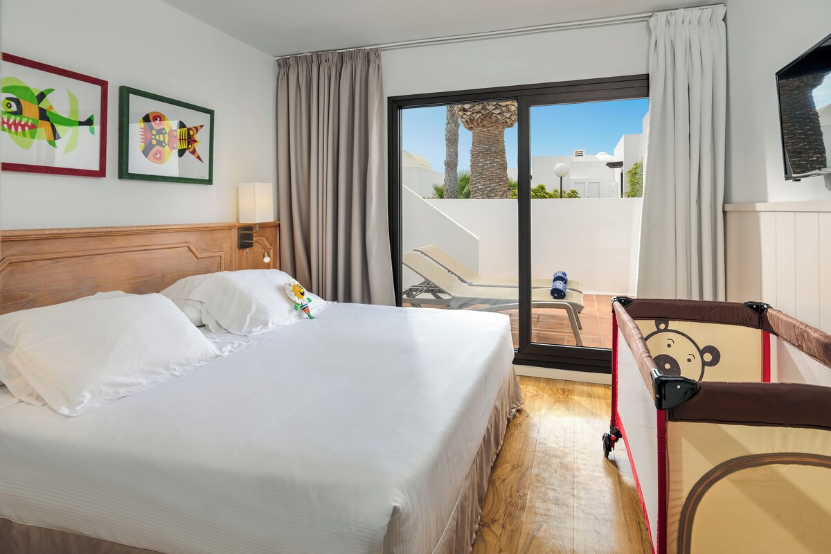 Habitación de hotel con cama, ventana con vista a palmeras y balcón con una silla.