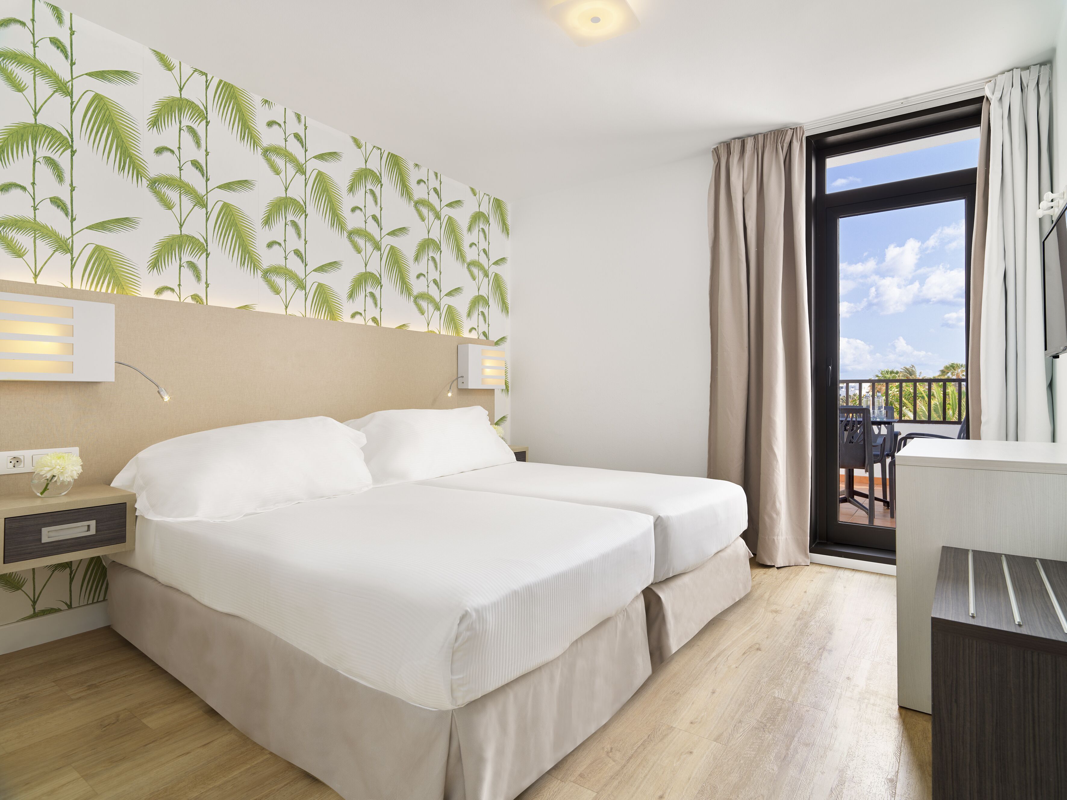 Habitación de hotel con cama, escritorio, balcón con vistas y una pared con estampado de hojas de palmera.