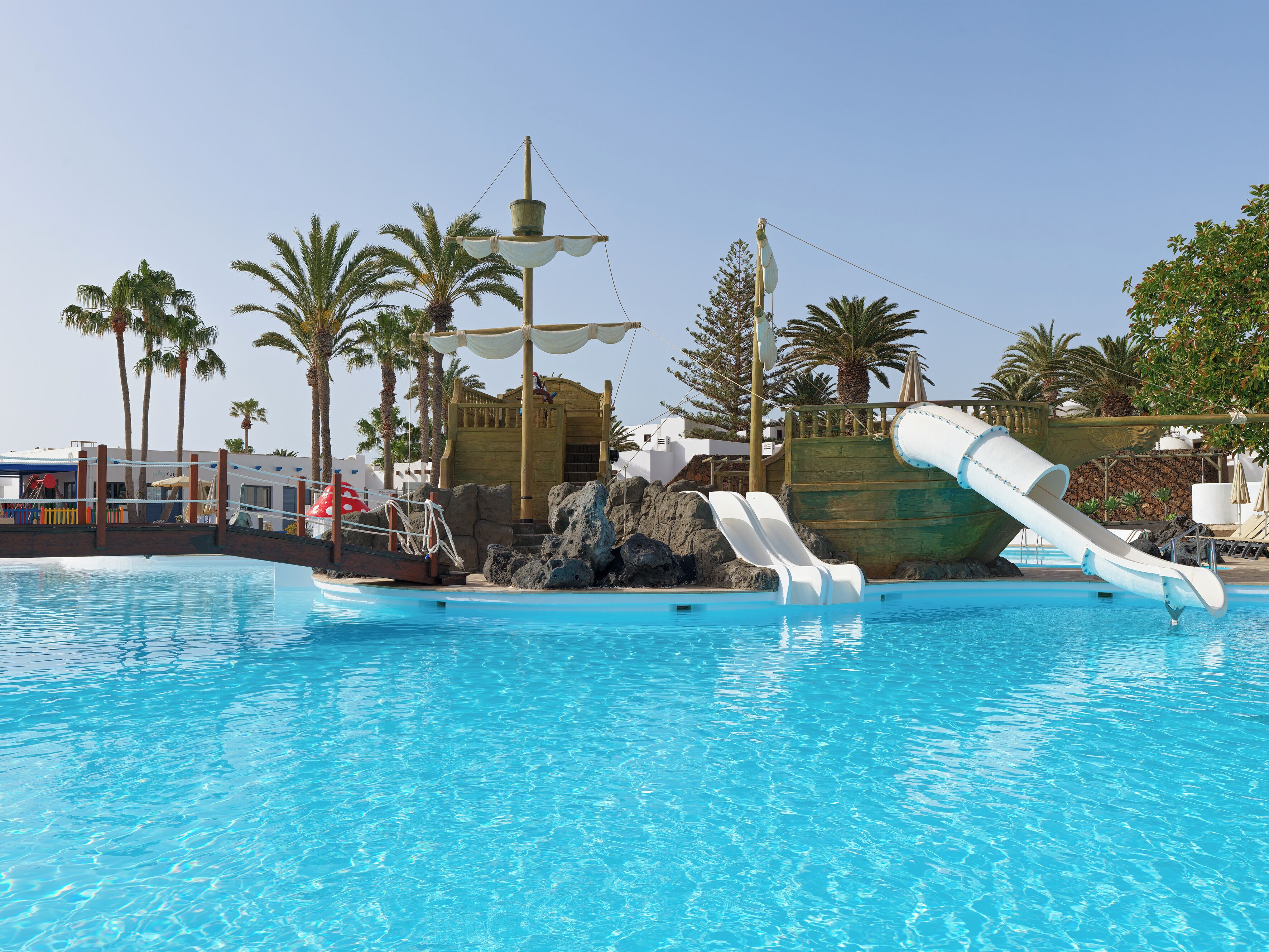 En swimmingpool med en stor rutschkana formad som ett skepp, palmer och en träkonstruktion.
