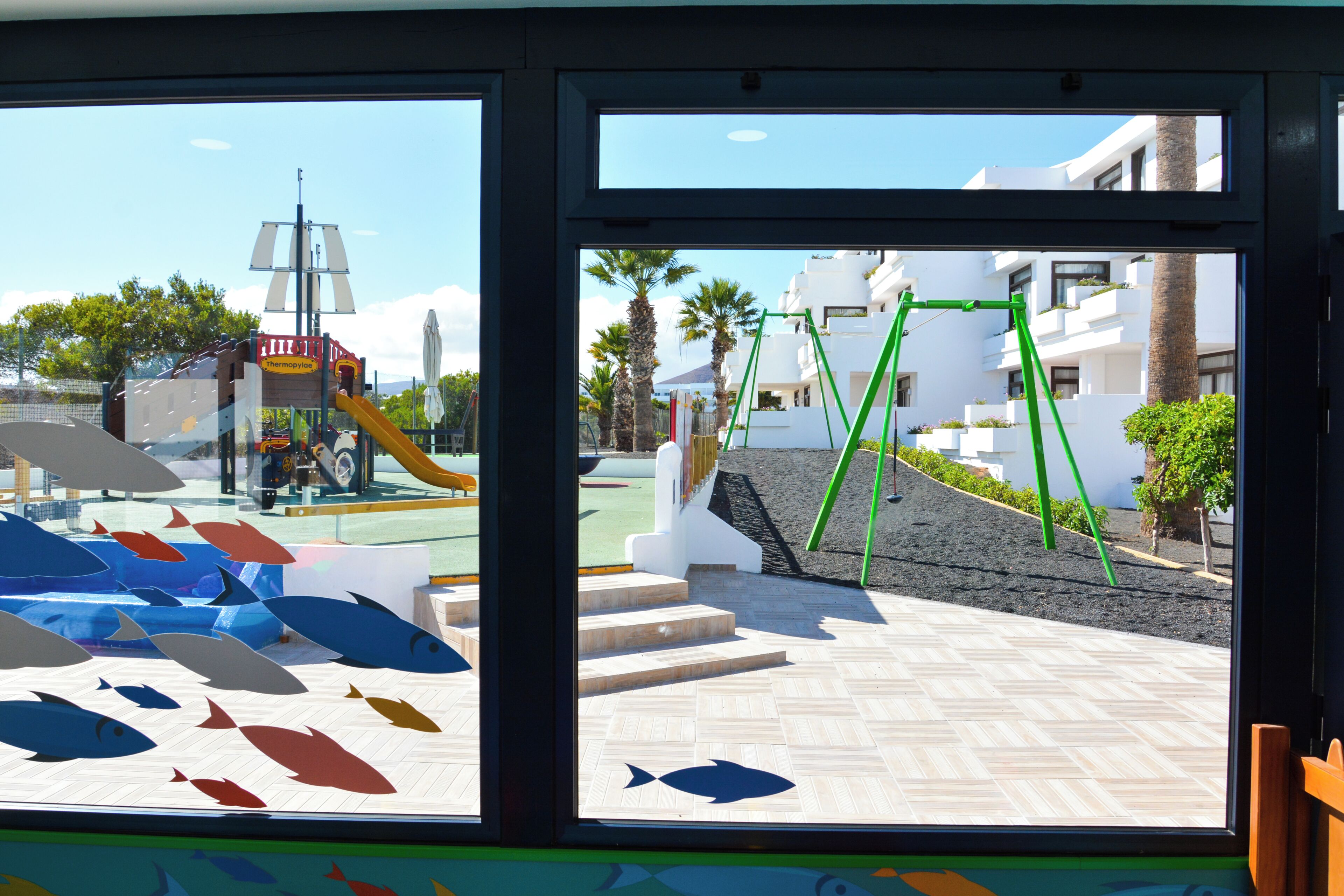 Vista a través de una ventana de un parque infantil con tobogán y estructuras de escalada, una zona empavedada con escalones y un edificio con balcones y palmeras.