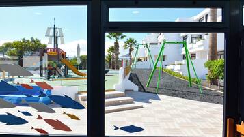 Vista a través de una ventana de un parque infantil con tobogán y estructuras de escalada, una zona empavedada con escalones y un edificio con balcones y palmeras.