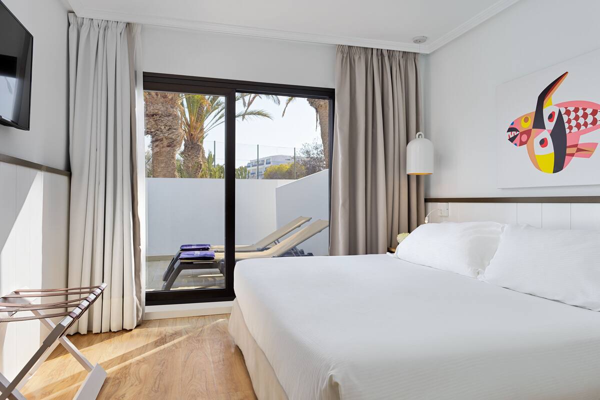 Une chambre d’hôtel avec un lit, une télévision, une fenêtre avec des rideaux, une chaise et un tableau au mur.