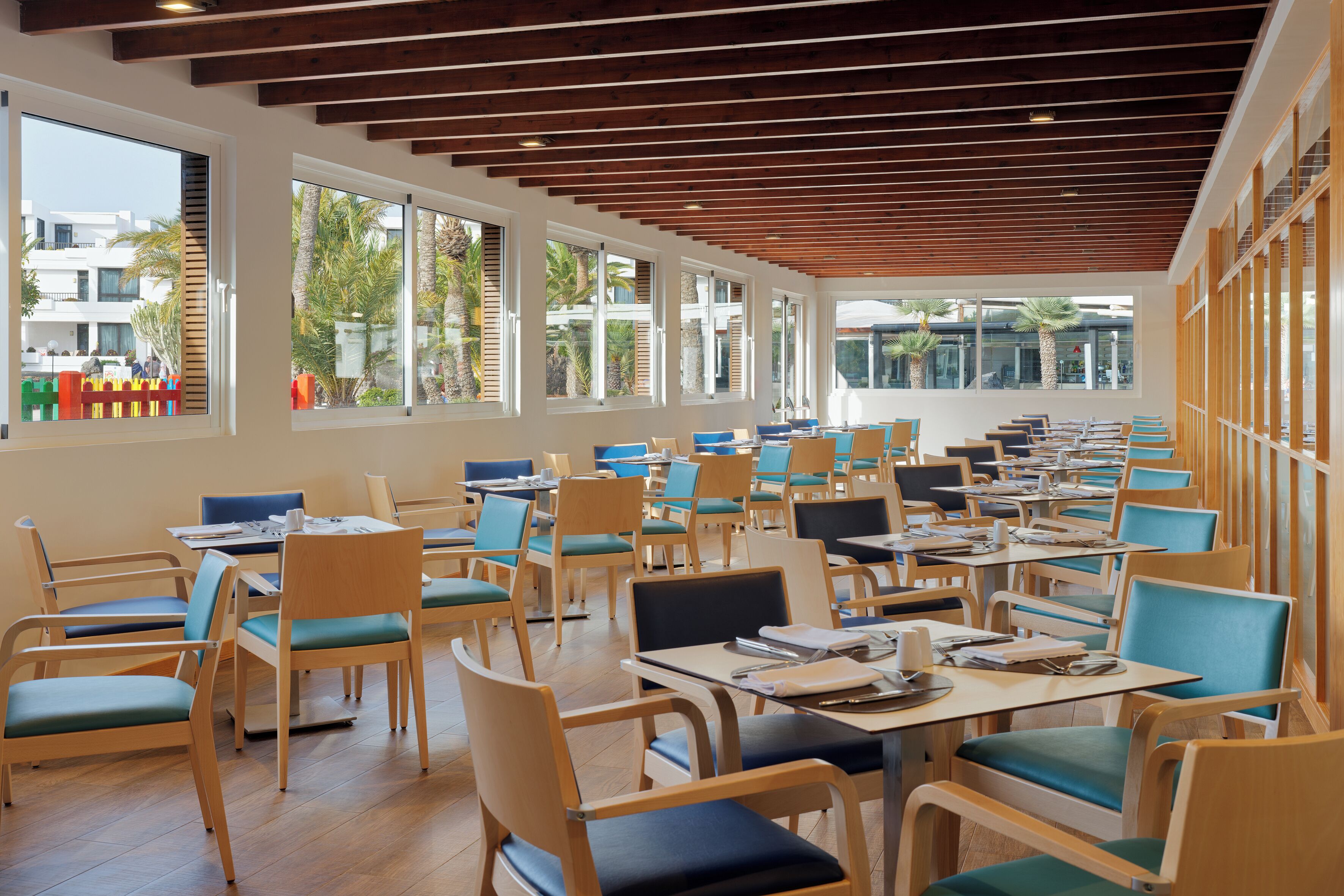 A spacious indoor dining area with multiple tables and chairs, large windows, and a wooden ceiling.