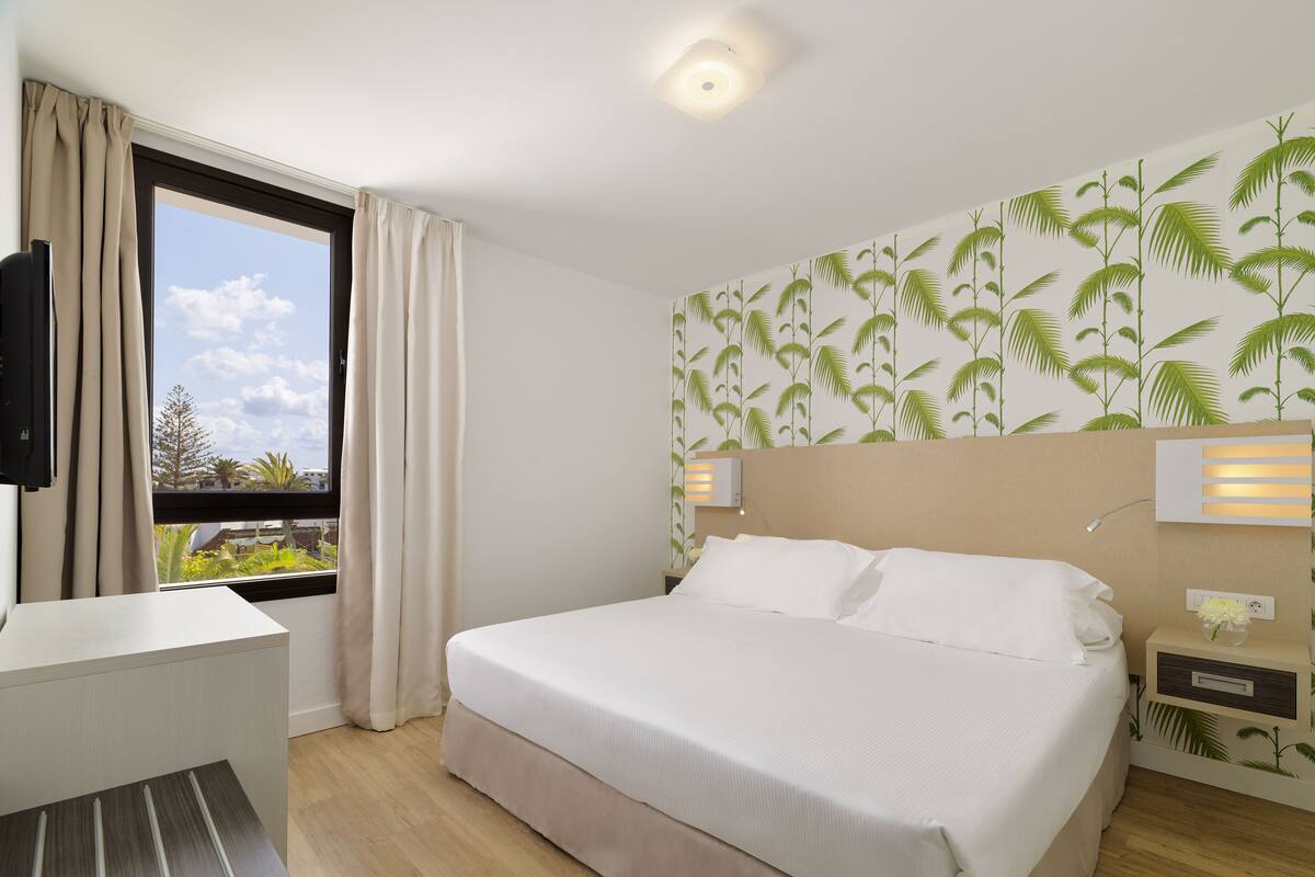 Une chambre d’hôtel comprenant un lit, des tables de chevet, un bureau, une fenêtre avec des rideaux et un mur orné d’un motif de palmiers.