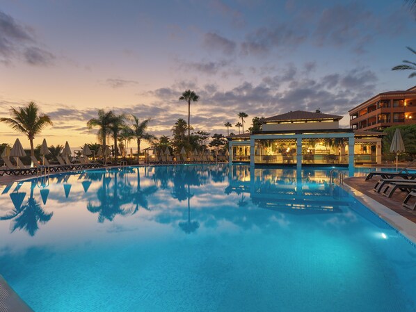 A swimming pool with lounge chairs and umbrellas, a bar area, and a building with a balcony. - H10 Costa Adeje Palace (Adeje)