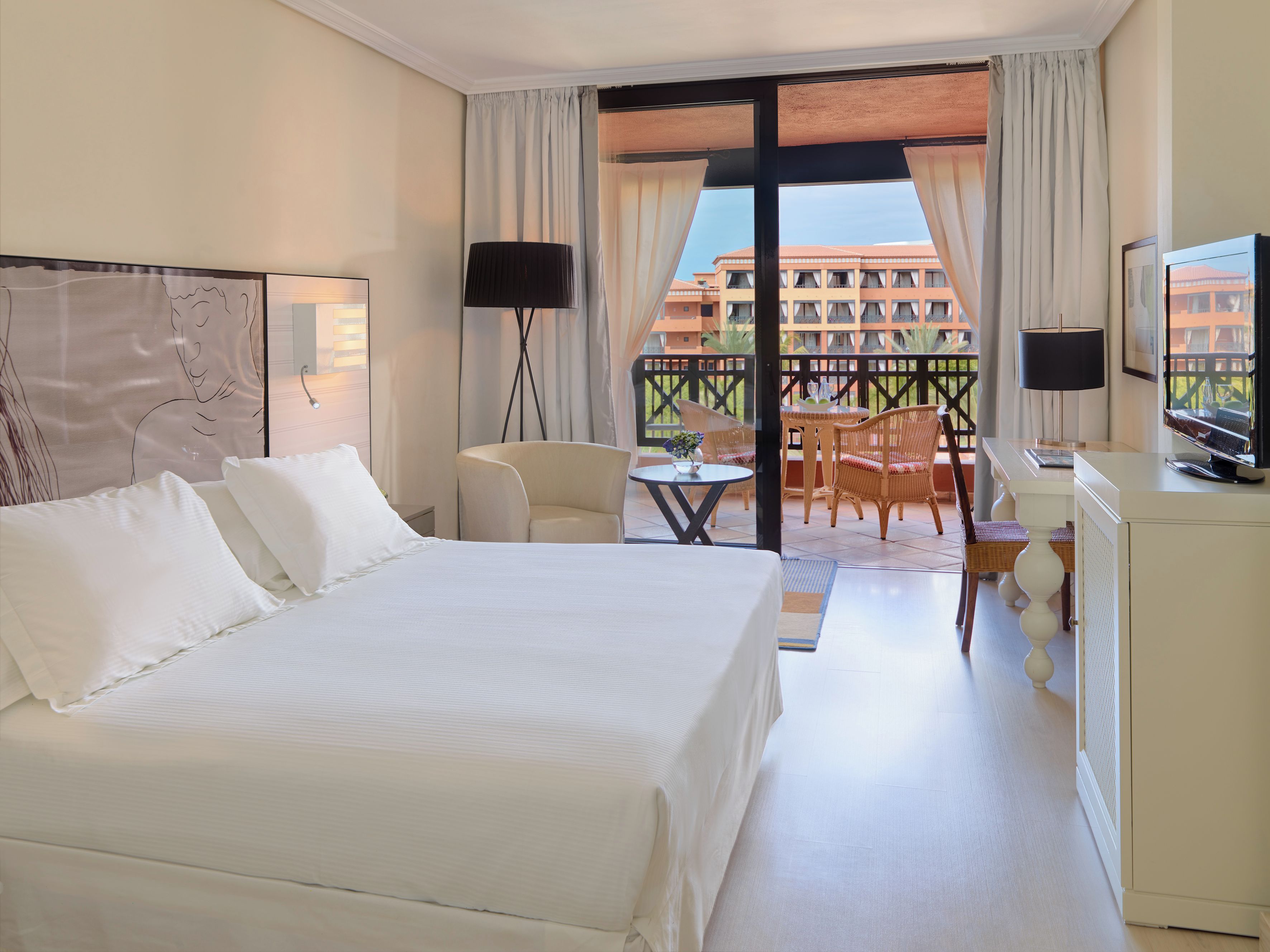A hotel room with a large bed, a desk with a TV, a chair, a small table with a vase, and a balcony with a view of a building.