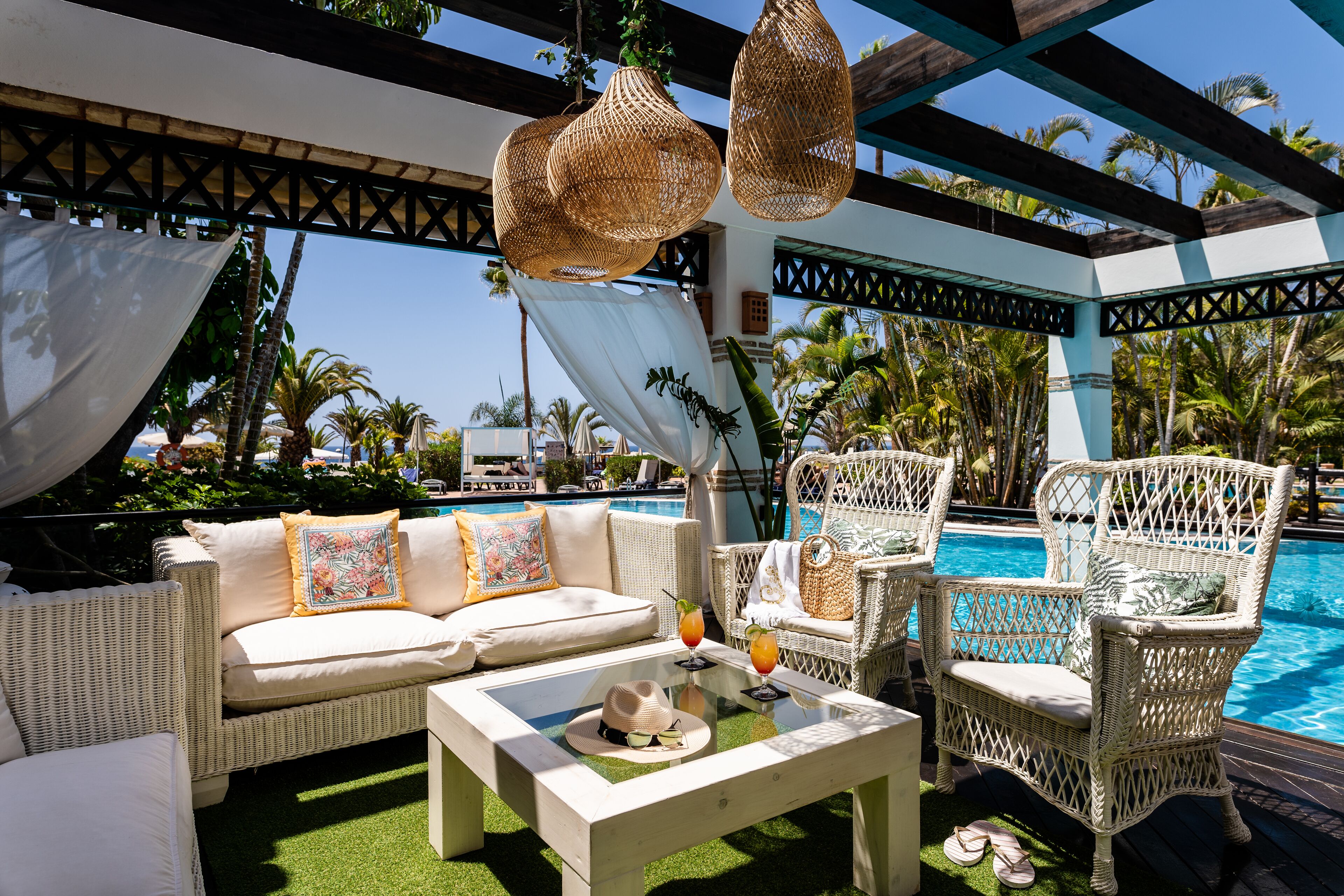 Un espace aménagé au bord de la piscine, avec des meubles en osier, un canapé blanc et une table en verre sur laquelle sont posés des boissons et un chapeau.